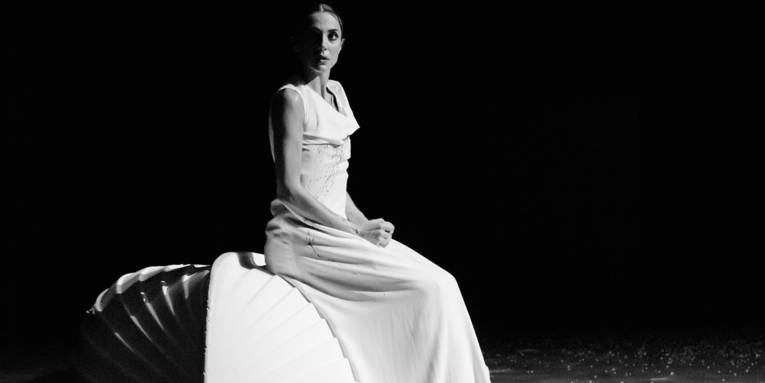 Auf einem schwarz-weiß Foto sitzt eine weiblich gelesene Person seitlich, in einem langen weißen Kleid, auf einem umgedrehten Boot und schaut über die Schulter nach links.